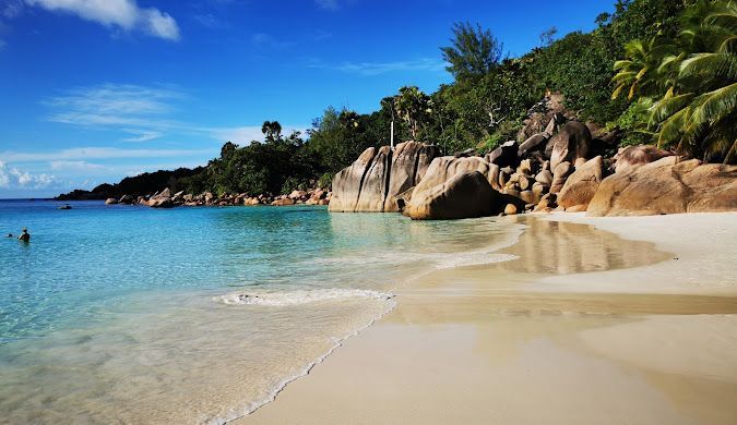 Onde leggere lambiscono una spiaggia di sabbia bianca accanto ad acqua turchese limpida, con grandi massi di granito e una lussureggiante collina verde sotto un cielo blu.