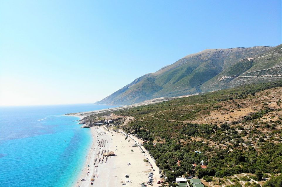 An aerial view of a long sandy beach with turquoise water next to a large green mountain under a clear blue sky.