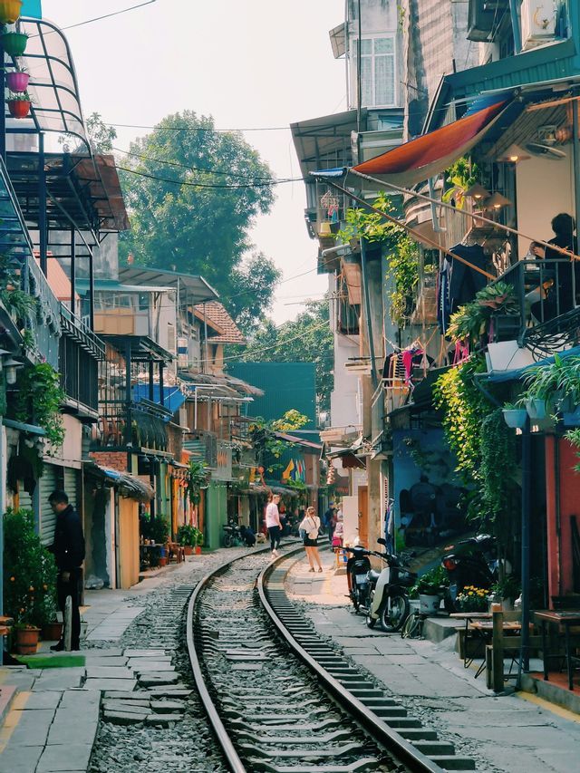 Binari del treno curvano attraverso una stretta via urbana fiancheggiata da edifici a più piani, piante in vaso e persone.