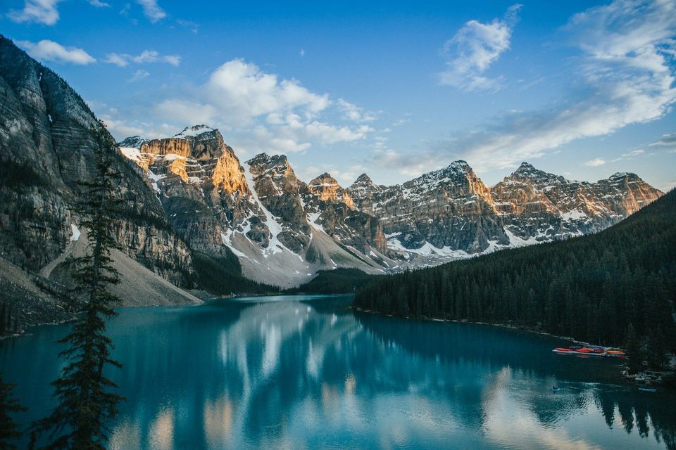 Sunlight hits the peaks of snow-capped mountains which reflect in a calm, turquoise lake surrounded by a pine forest.