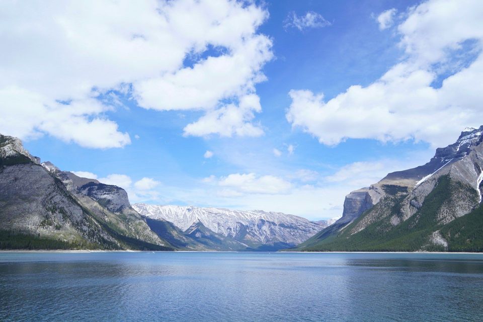 A wide, calm lake is flanked by steep, forested mountains under a blue sky with fluffy white clouds.