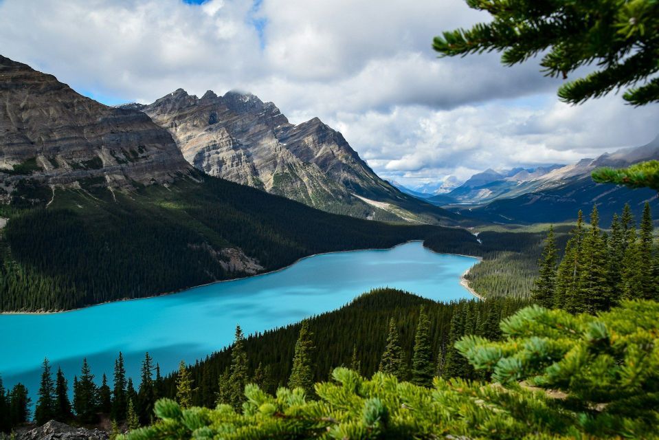 An elevated view of a bright turquoise lake surrounded by dense pine forests and rocky mountains under a cloudy sky.