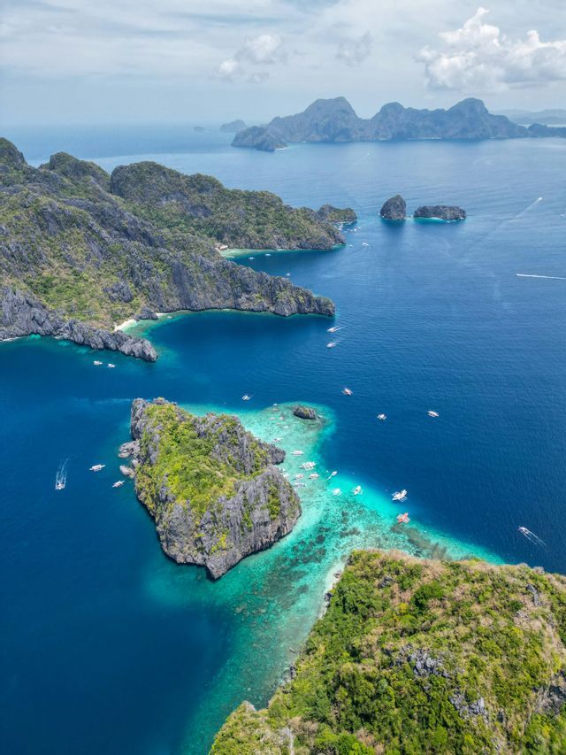 Eine Luftaufnahme von üppigen, felsigen Inseln in einem türkisfarbenen und tiefblauen Meer, mit vielen kleinen Booten, die über das Wasser verteilt sind.