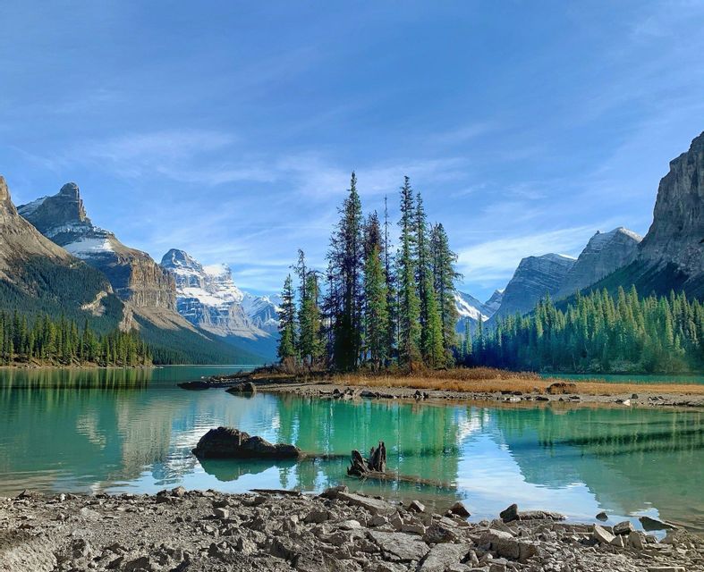 A small island with pine trees sits in a calm turquoise lake, surrounded by forests and snow-capped mountains.