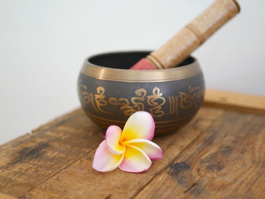 Eine tibetische Klangschale mit goldener Schrift und einem Klöppel steht neben einer Frangipani-Blüte auf einer rustikalen Holzoberfläche.
