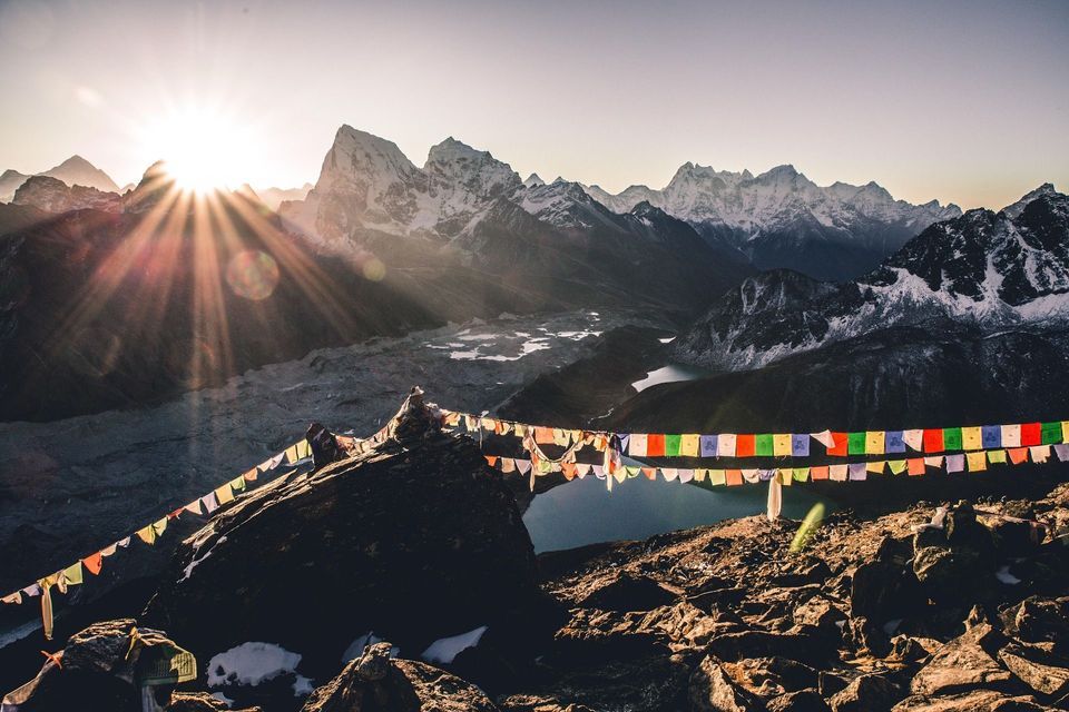 Bunte Gebetsfahnen wehen über einer felsigen Landschaft, während die Sonne hinter schneebedeckten Bergen aufgeht und ein Tal mit einem See darunter liegt.