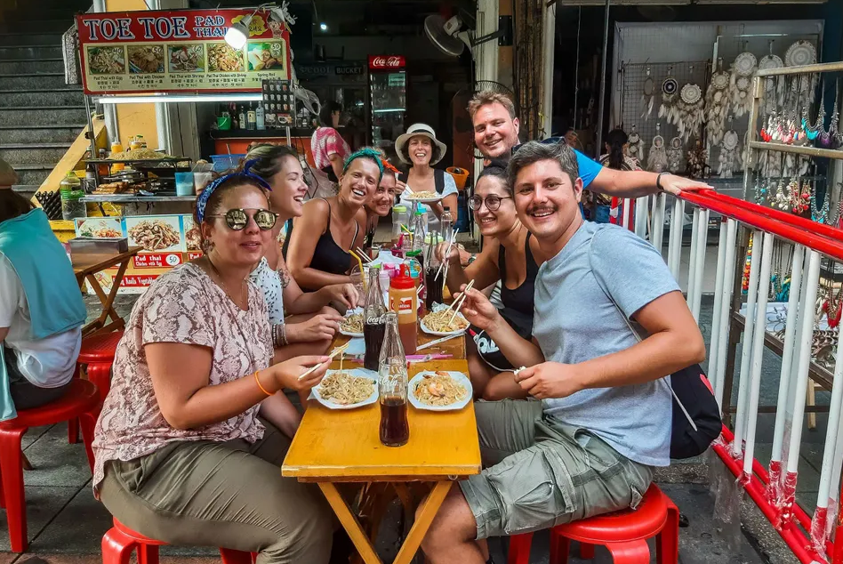 Eine WeRoad-Gruppe, die lächelnd an einem langen Tisch neben einem Streetfood-Stand Pad Thai mit Essstäbchen isst.