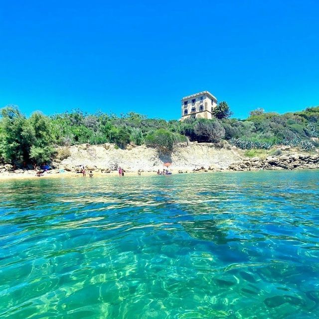 Acqua cristallina turchese in primo piano, con una spiaggia sabbiosa e una torre di pietra su una collina verde sullo sfondo sotto un cielo sereno.