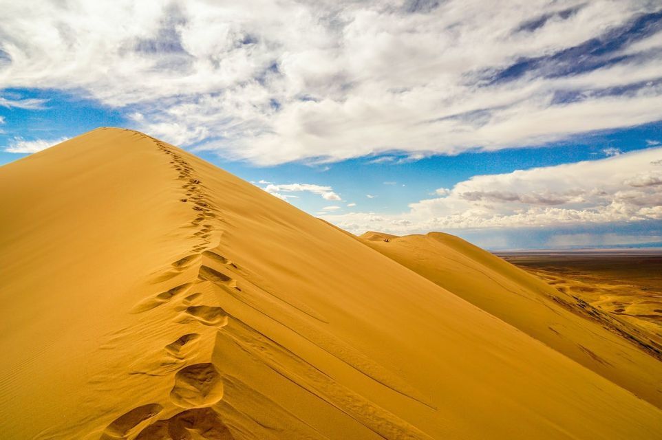 Une trace de pas monte au sommet d'une grande dune de sable doré sous un ciel bleu partiellement nuageux.