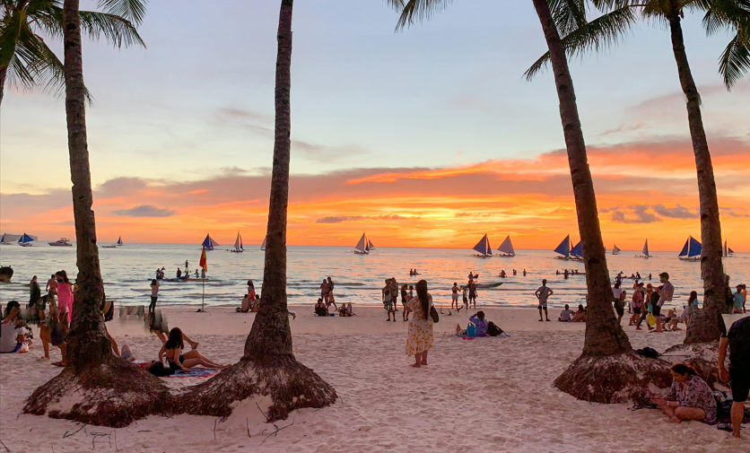 Menschen entspannen sich an einem Sandstrand mit Segelbooten auf dem Meer während eines lebhaften Sonnenuntergangs, umrahmt von Palmen.