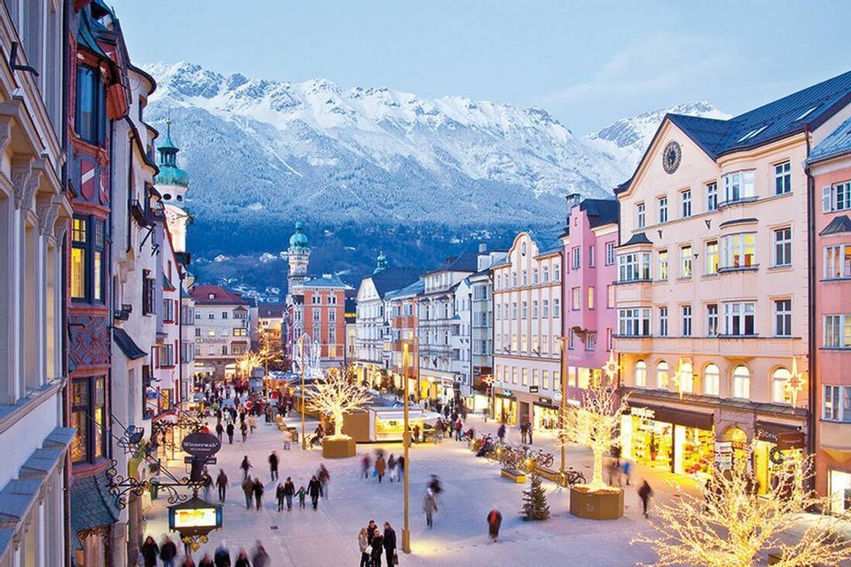 Una vivace via pedonale in una città in stile europeo al crepuscolo, con edifici colorati e grandi montagne innevate sullo sfondo.