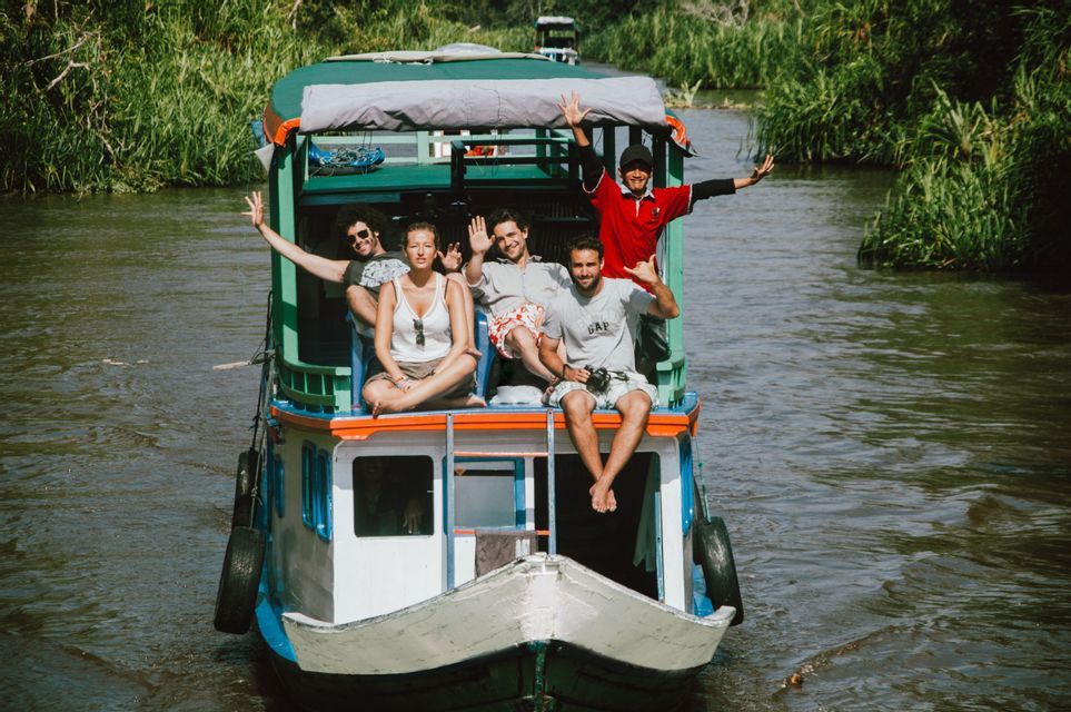 Un grupo de WeRoad sonríe y saluda desde la cubierta superior de un barco en un río rodeado de vegetación.