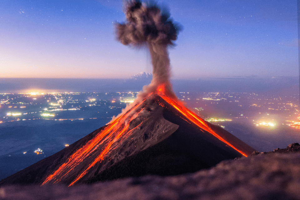 Ein nachts ausbrechender Vulkan, mit Flüssen aus geschmolzener Lava, die seine Hänge hinabfließen, und Rauch, der in einen sternenklaren Himmel über einer Stadt aufsteigt.