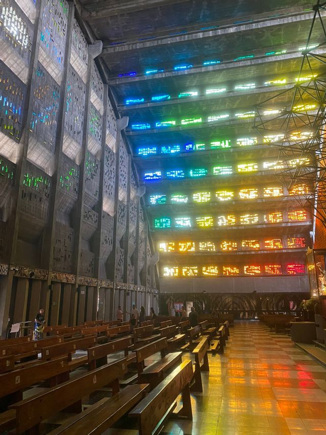 Das Innere einer modernen Betonkirche, in der farbiges Licht von Buntglasfenstern die Holzbänke beleuchtet.