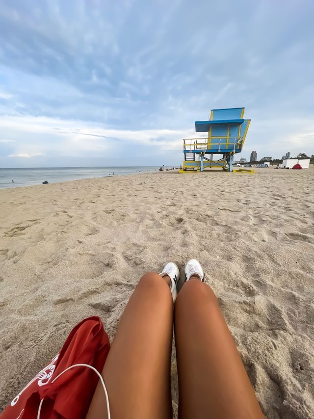 A first-person perspective of a person sitting on a sandy beach, with a blue and yellow lifeguard tower in the distance.