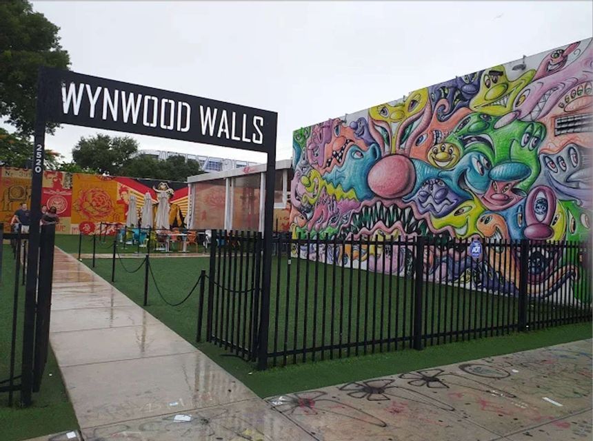 The entrance to Wynwood Walls on a wet day, featuring a large black sign and colorful murals behind a fence.