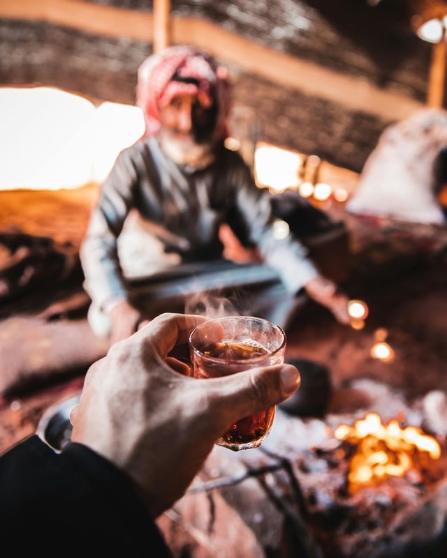 Una persona durante un viaggio di gruppo WeRoad tiene in mano un bicchiere di tè fumante accanto a un fuoco da campo all'interno di una tenda rustica.