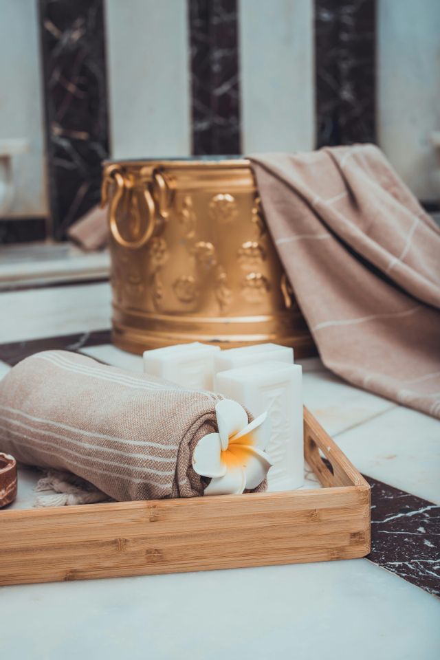 Un asciugamano arrotolato, saponi e un fiore di plumeria disposti su un vassoio di legno, con un secchio dorato in un ambiente da spa.