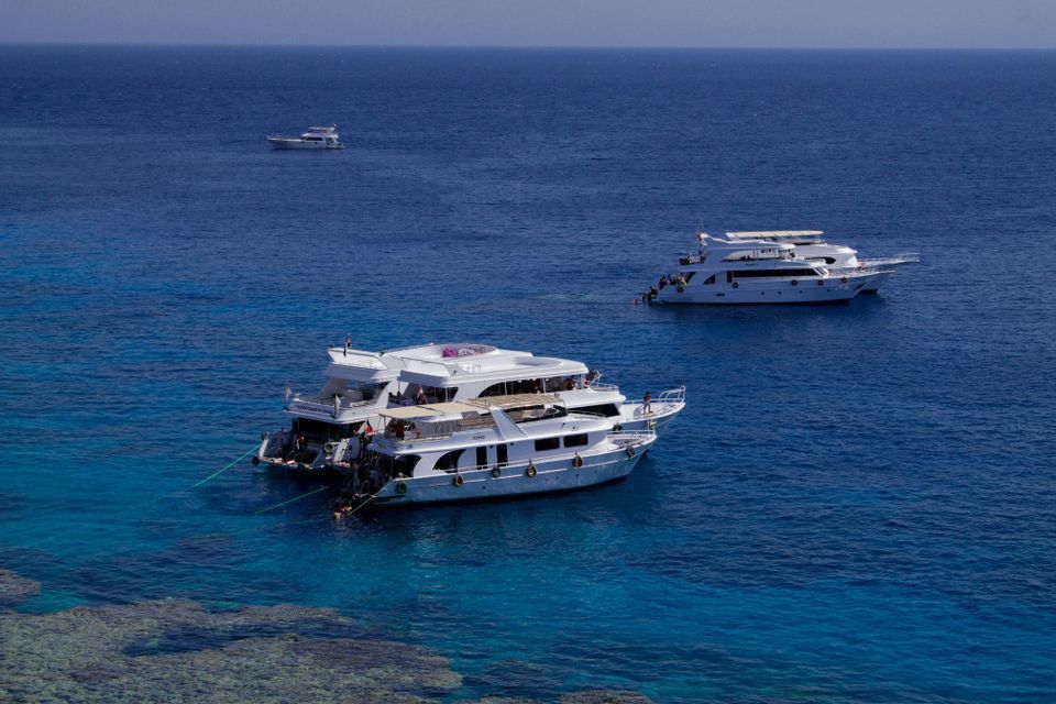 Diversi yacht bianchi di un gruppo WeRoad sono ancorati in acque turchesi sopra una barriera corallina visibile, sotto un cielo azzurro e limpido.