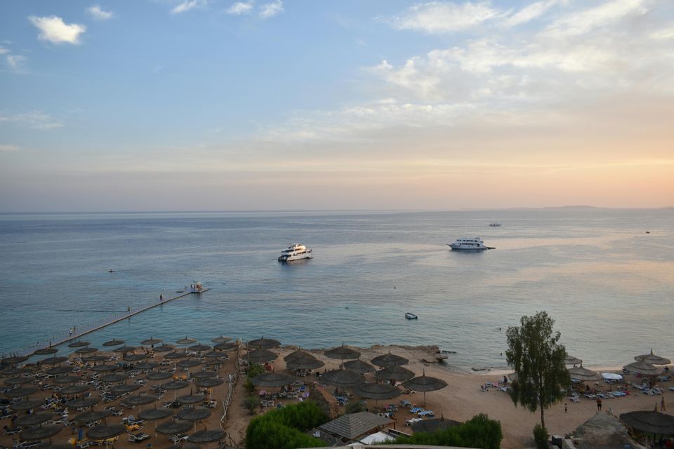 Una vista dall'alto di una spiaggia sabbiosa con ombrelloni di paglia e sedie a sdraio, con diversi yacht sul mare calmo al tramonto.