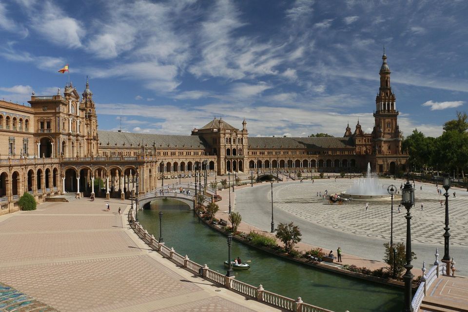 Un edificio semicircolare ornato con torri che si affaccia su un'ampia piazza con un canale e una fontana, sotto un cielo azzurro nuvoloso.