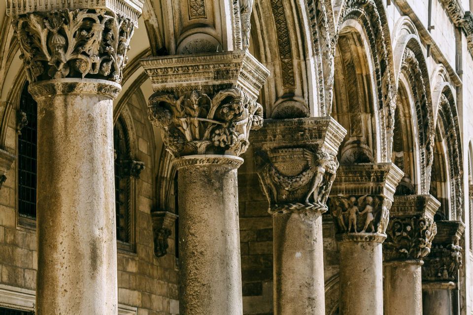 Primo piano di colonne in pietra consumata con capitelli finemente scolpiti, parte di una facciata ad arco di un edificio storico.