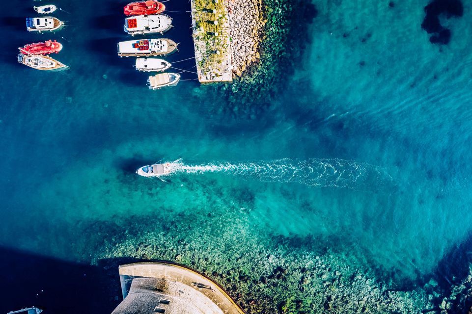 Una veduta aerea, dall'alto, di un motoscafo che crea una scia in acqua turchese, passando accanto a un molo di pietra con altre barche ormeggiate.