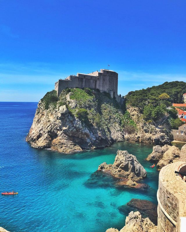 Una fortezza in pietra si erge su una scogliera rocciosa e verde che domina un mare turchese con due kayak rossi che remano nell'acqua sottostante.