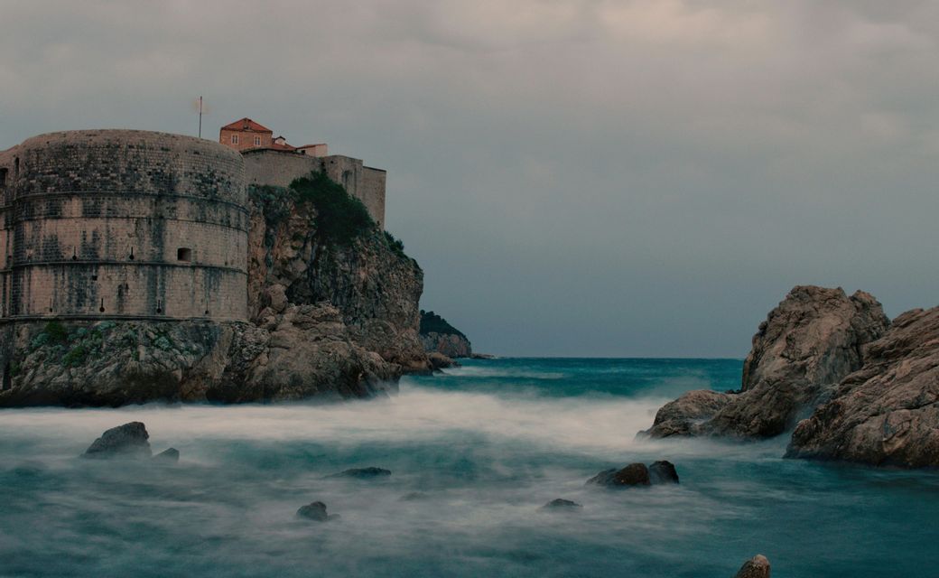 Una fortezza in pietra su una scogliera rocciosa, con onde a lunga esposizione che creano un effetto nebbioso sul mare sottostante sotto un cielo nuvoloso.