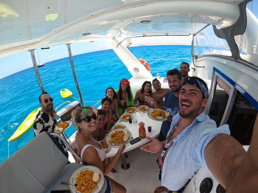 Eine WeRoad-Reisegruppe lächelt für ein Selfie beim gemeinsamen Essen auf einem Boot inmitten türkisfarbenen Meeres.