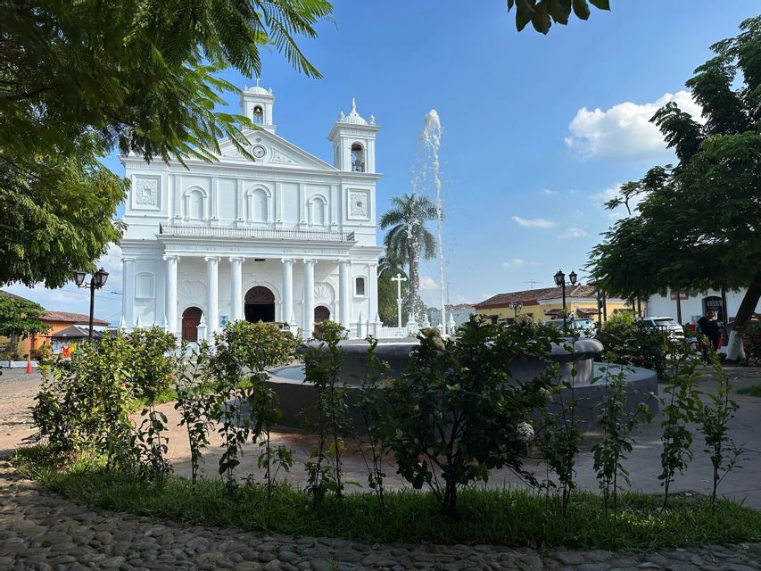 Eine weiße Kolonialkirche mit Säulen und Glockentürmen steht hinter einem Brunnen auf einem sonnigen Stadtplatz, von einem Park aus gesehen.