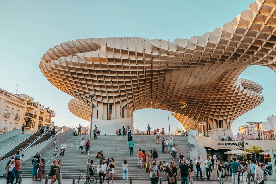 Una multitud de personas en una escalera ancha bajo un gran dosel de madera contemporáneo con diseño de celosía, contra un cielo despejado.