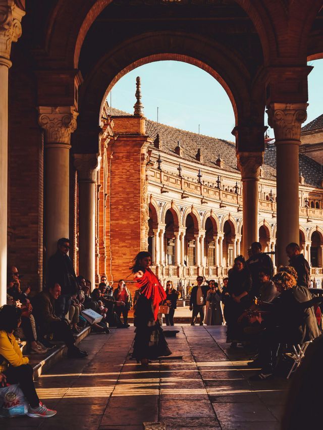 Una bailaora de flamenco con mantón rojo actúa para un público en una plaza soleada y con arcos, de arquitectura de ladrillo.