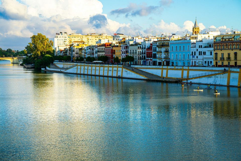 Un grupo de personas en tablas de paddle surf flota en un río ancho junto a un paseo marítimo con edificios coloridos bajo un cielo parcialmente nublado.