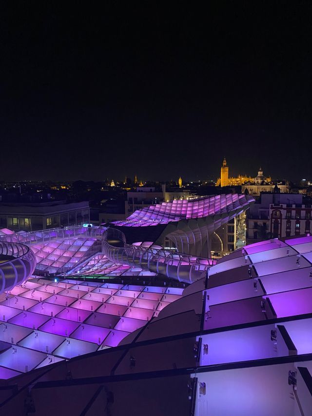 Una estructura moderna con patrón de cuadrícula, iluminada con luces moradas, domina el horizonte de una ciudad por la noche.