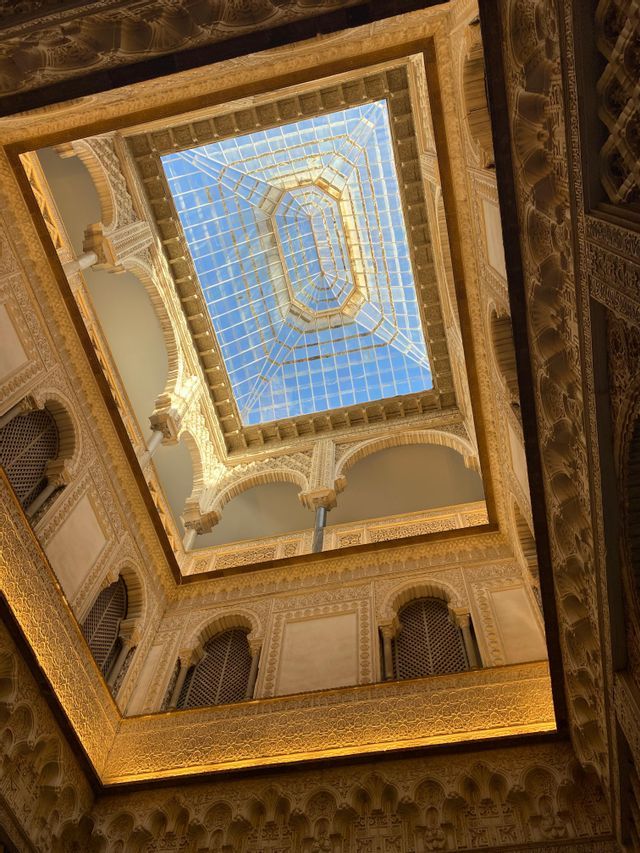 Una vista en contrapicado, mirando hacia arriba a través de un patio ornamentado con arcos tallados, hacia un techo de cristal que deja ver un cielo azul.