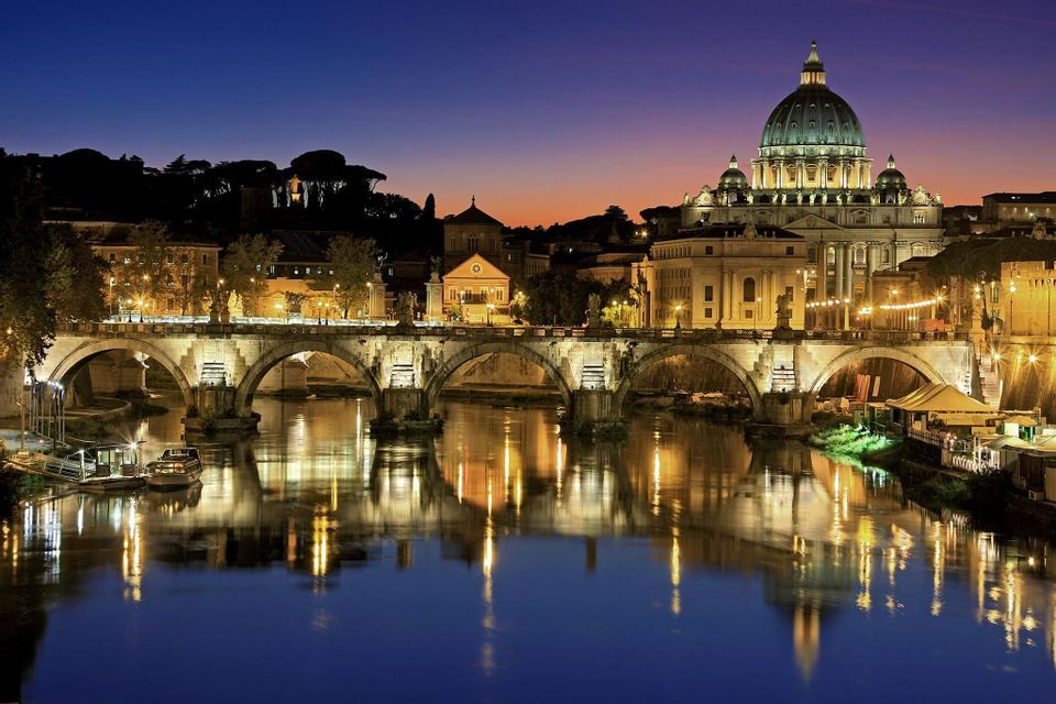 Un ponte di pietra illuminato e una grande basilica a cupola riflessi in un fiume al crepuscolo contro un cielo blu scuro.
