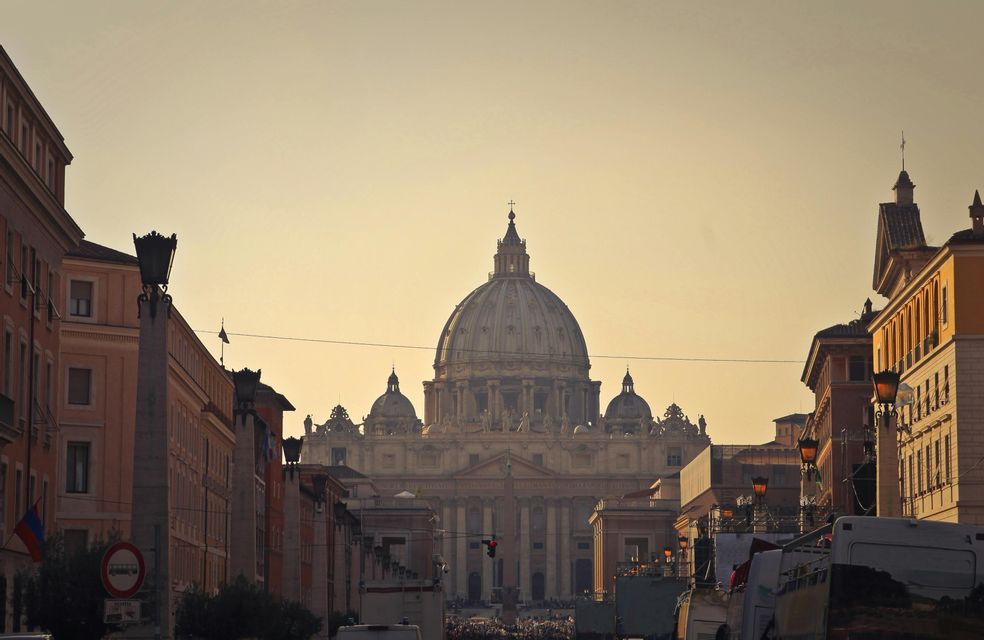 A large domed basilica at the end of a street, with crowds in the distance and buildings on either side, bathed in warm sunset light.
