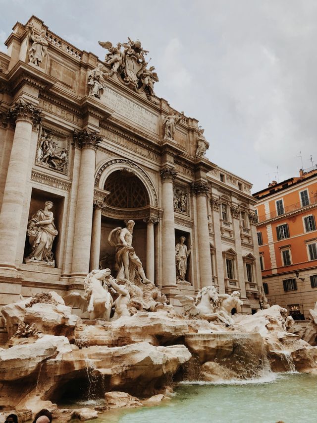 Una fontana barocca riccamente ornata, con grandi statue di figure e cavalli, sotto un cielo nuvoloso.