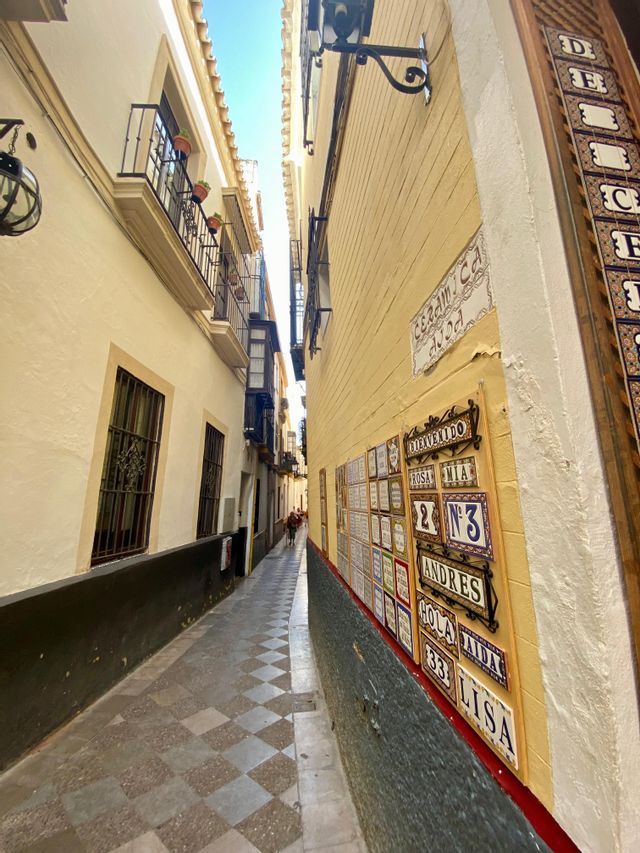 Un callejón estrecho con paredes amarillas, un pavimento de damero y azulejos cerámicos decorativos montados en un lado bajo un cielo azul.