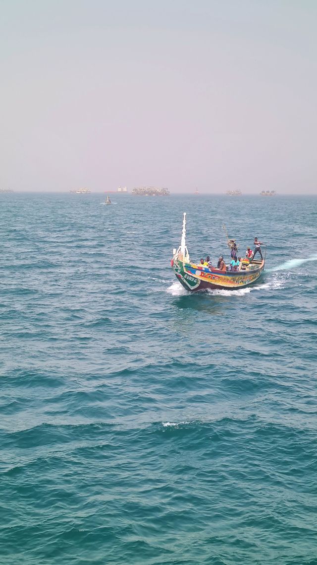 Eine Gruppe von Menschen auf einem farbenfrohen Holzboot segelt über ein unruhiges blaues Meer, mit weiteren Booten in der diesigen Ferne.