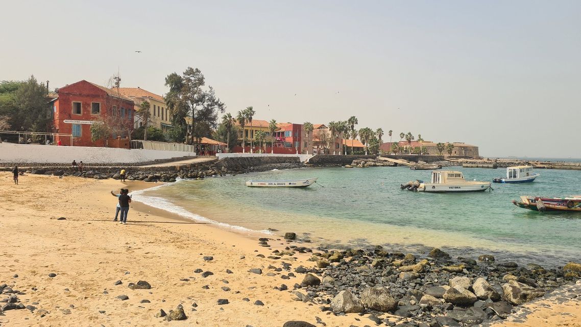 Ein sandiger, felsiger Strand an einer ruhigen Bucht mit vertäuten Booten, bunten Gebäuden und Palmen entlang der Uferpromenade.