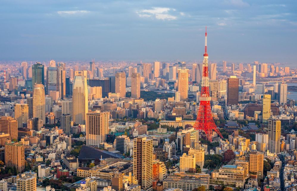 An aerial view of a dense cityscape with a prominent red and white tower illuminated by the warm light of sunset.