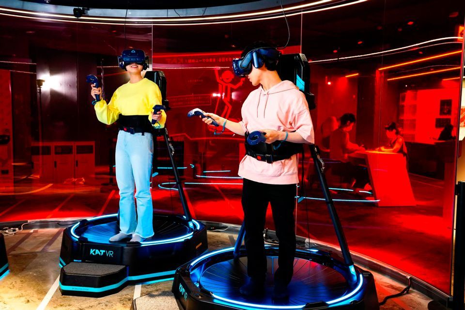 Due persone con visori VR e controller si trovano su piattaforme di gioco circolari in una stanza con illuminazione rossa e blu.