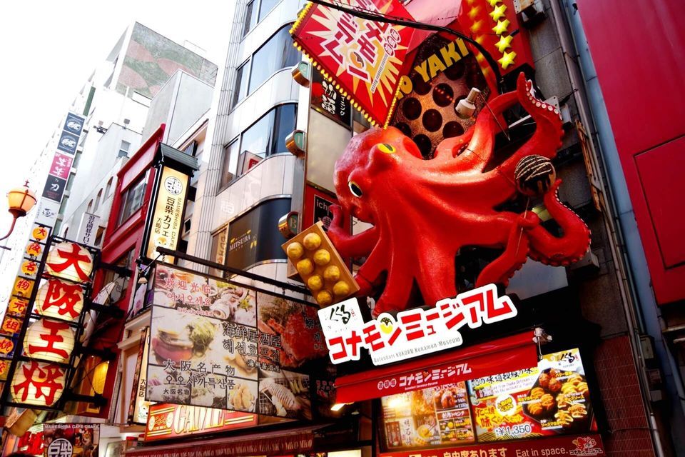 Una scultura gigante di polpo rosso che tiene takoyaki è montata su un edificio sopra una trafficata strada giapponese con molte insegne colorate.