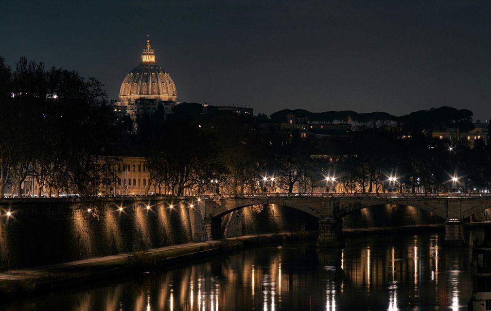 Una cúpula iluminada se eleva sobre el horizonte de una ciudad por la noche, con un puente de piedra y farolas reflejándose en el río de abajo.