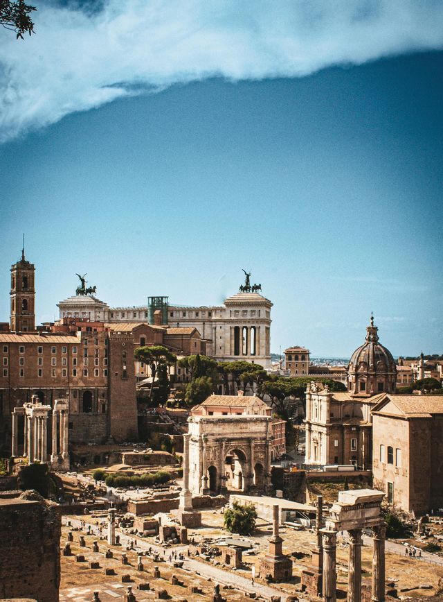 A high-angle view of ancient ruins, including arches and columns, with a historic city skyline under a blue sky.