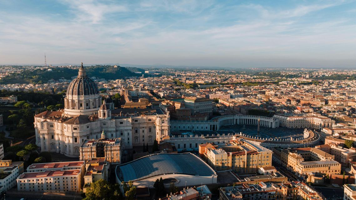 Eine Luftaufnahme einer prachtvollen Basilika mit einer großen Kuppel und einem weitläufigen Säulenplatz, die eine ausgedehnte Stadt bei Sonnenaufgang überblickt.