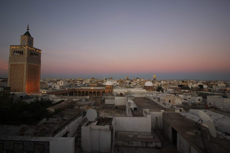 Un minaret illuminé domine un paysage urbain de bâtiments blancs et de toits plats sous un ciel rose et violet au crépuscule.