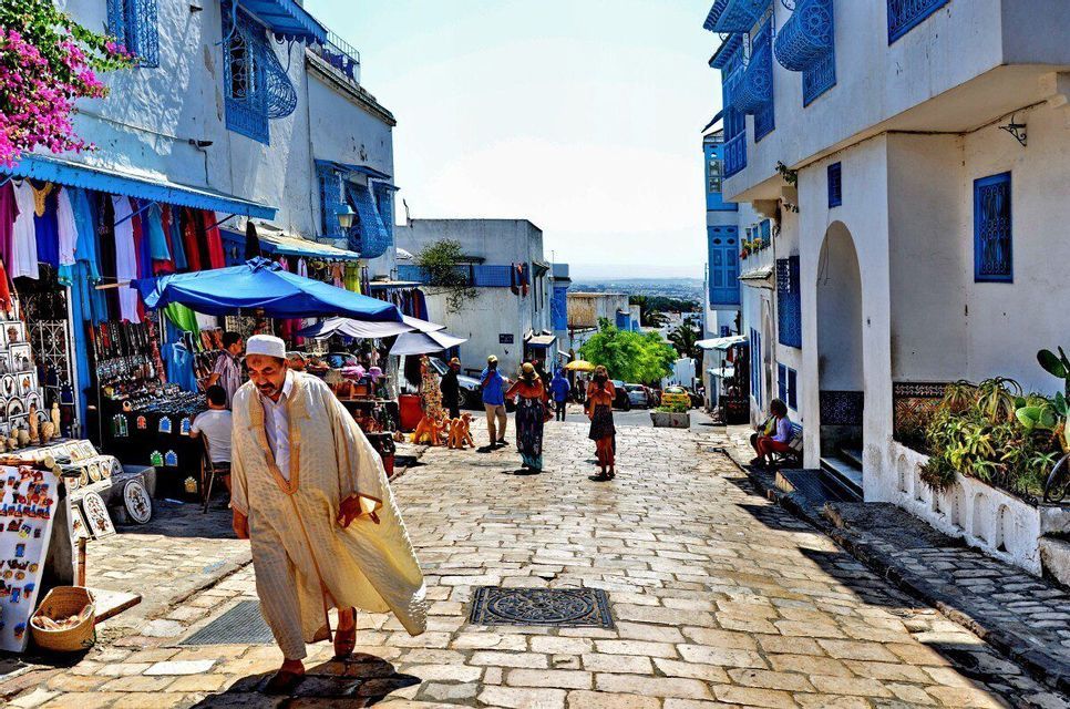 Un homme en tenue traditionnelle marche dans une rue pavée animée bordée d'étals de marché et de bâtiments blanchis à la chaux avec des garnitures bleues.